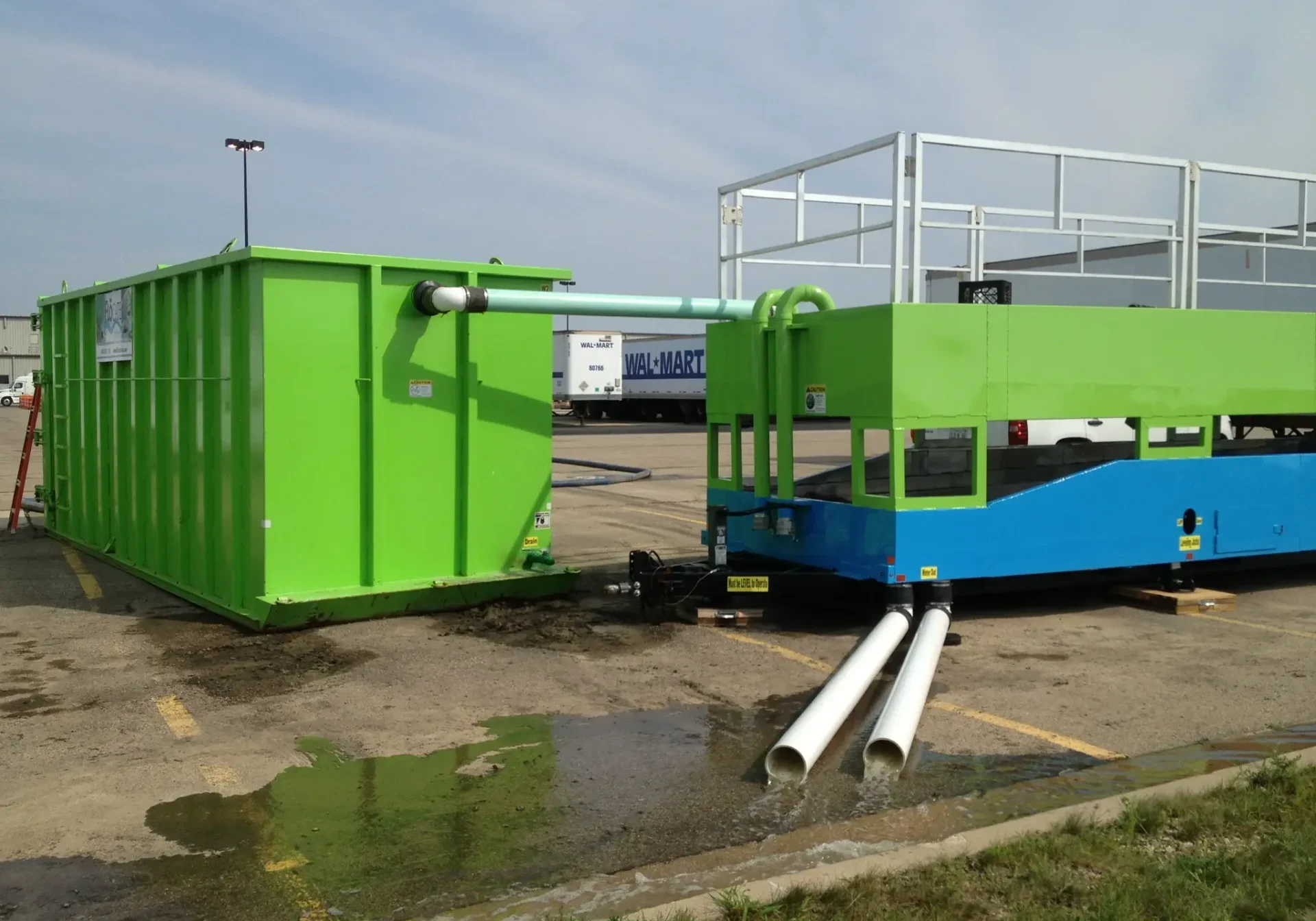 Industrial containers and pipes on a wet concrete surface near a parking area.