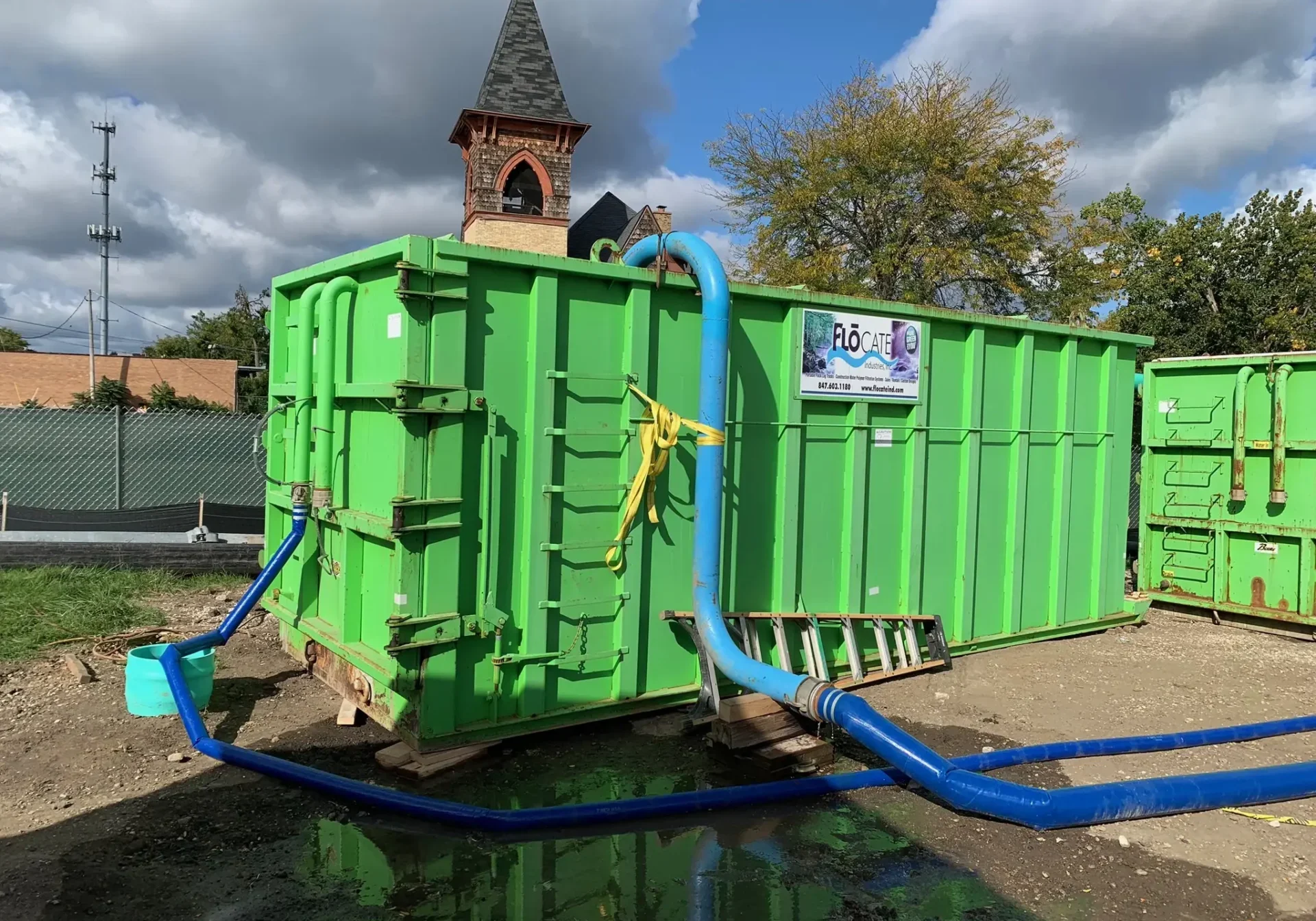 Large green container with hoses and pipes outdoors under a cloudy sky.
